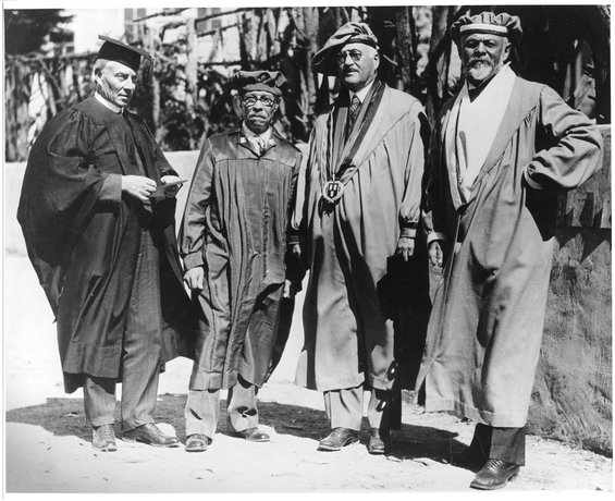 Black and white photo with four people in long academic robes. The one on the left is wearing a mortarboard, and the three on the right are wearing tams (velvety, beret-like caps).