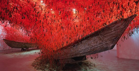 An installation at the 2017 Venice Biennale by Chiharu Shiota, two long, slim boats of distressed wood angled up from the ground. Rising out from the boats is a complex red web, forming a cloud above them from which dangle hundreds of metal keys.