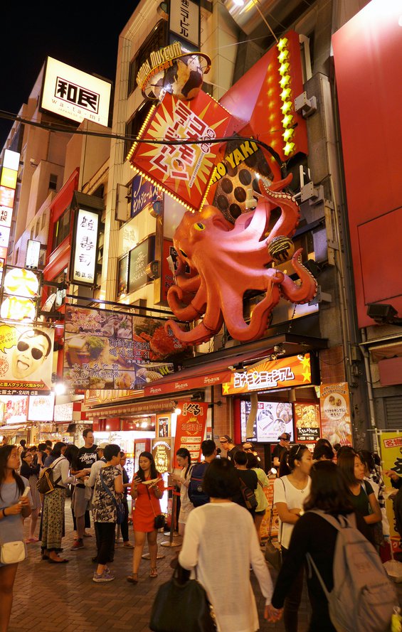 Shoppers walking the night market on Dotonbori street in Osaka below a large 3D pink octopus sign.