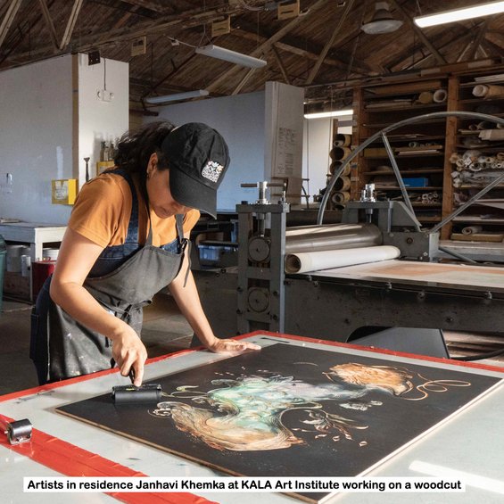 An artist applies black ink to a large woodcut with carved lines and shapes.