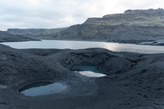 Dark grey Icelandic glaciers with two almost circular pools of water in the foreground and a long stretch of water in the middleground.