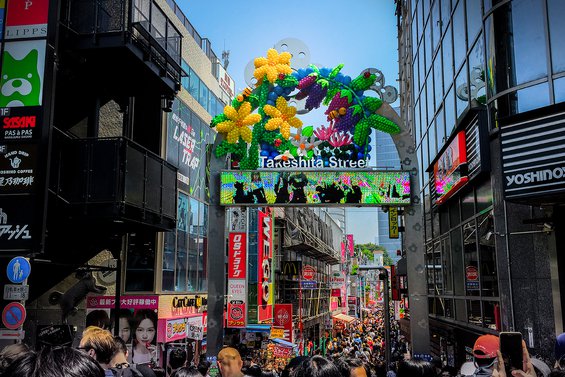 A metal arch, adorned with  yellow flowers, vines and grapes, between tall buildings in the Harajuku shopping and entertainment district in Tokyo.