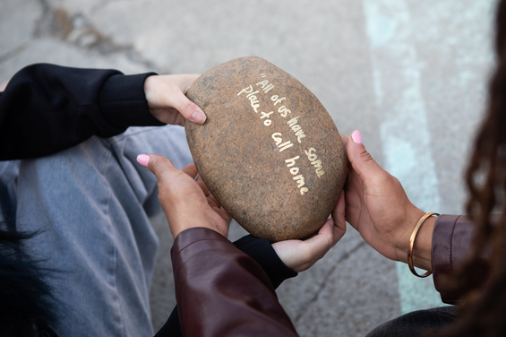 Hands holding a stone on which is written, "All of us have some place to call home."