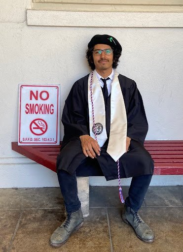 Wally Corona (BFA 2023) in tam and gown sitting on a bench
