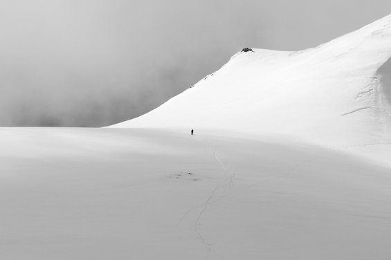 Black and white image of a white sky over a snowy expanse of ground with the minuscule figure of a man in its midst; a hill rises up from the ground to his right.